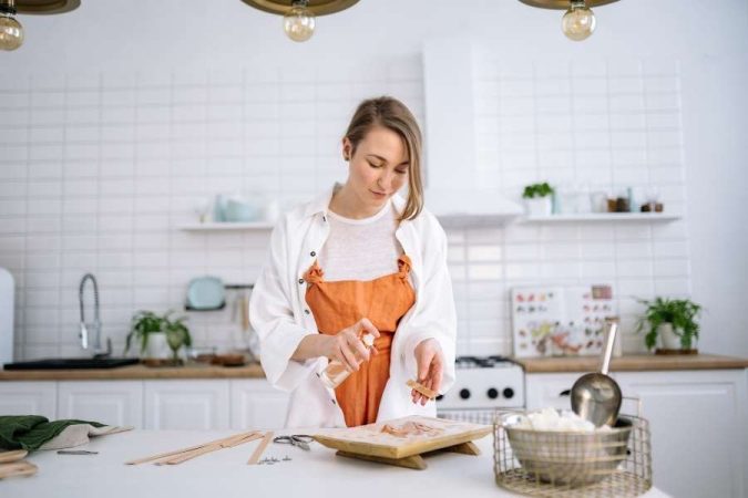 Femme en tablier orange préparant une recette sur une planche en bois dans une cuisine lumineuse, lors d'un cours de cuisine.