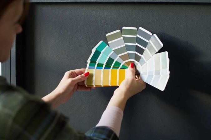Une femme aux ongles rouges compare deux éventails de palette de couleurs devant un mur gris foncé pour choisir une teinte de peinture.