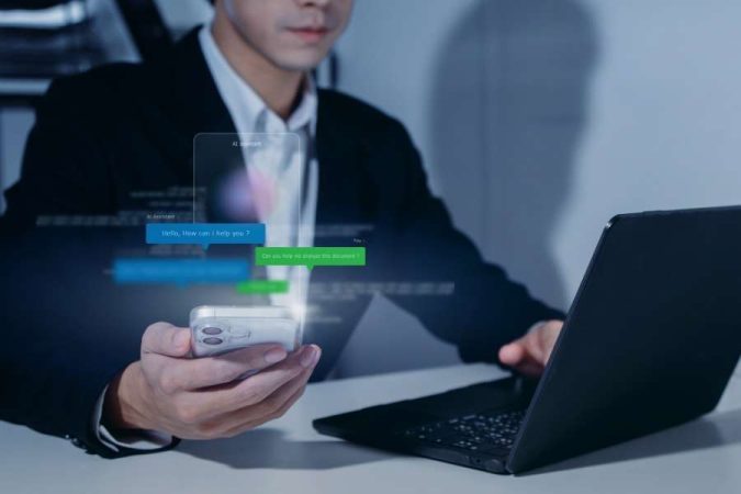 Un homme en costume utilise son smartphone d'où émerge une interface de messagerie instantanée virtuelle avec un assistant virtuel.