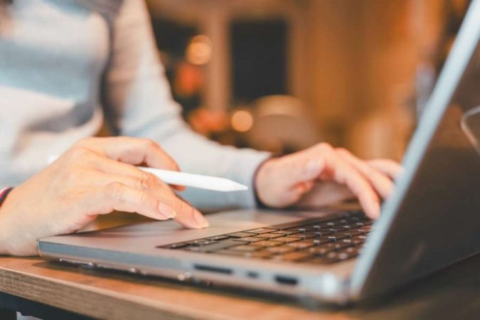 Gros plan sur les mains d'une personne écrivant sur le clavier d'un ordinateur portable tout en tenant un stylet blanc.