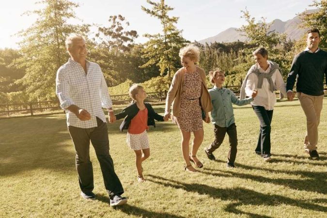 Une famille multigénérationnelle composée de grands-parents, parents et enfants se tient par la main en marchant joyeusement sur une pelouse verte lors d'une journée ensoleillée.